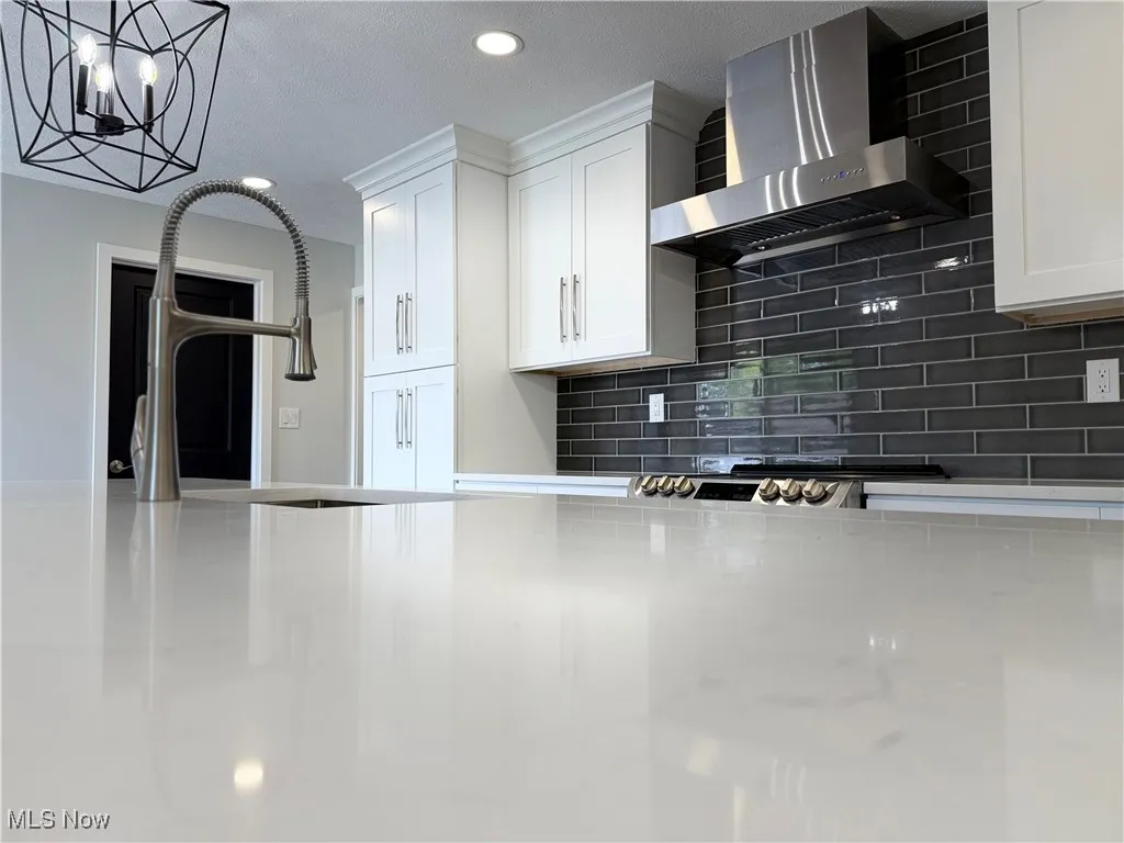Kitchen featuring wall chimney range hood, decorative backsplash, white cabinetry, recessed lighting, and a chandelier