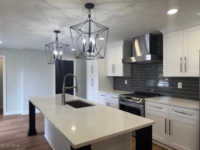 Kitchen with wall chimney exhaust hood, stainless steel range with electric cooktop, a center island with sink, decorative backsplash, and white cabinetry