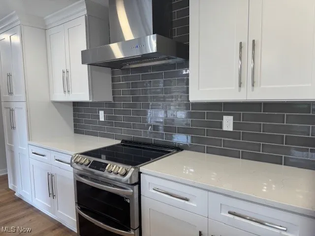 Kitchen with double oven range, wall chimney exhaust hood, white cabinetry, and light stone counters