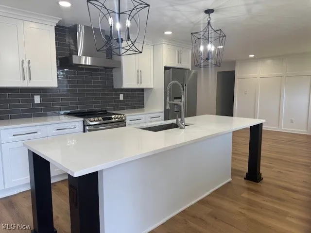 Kitchen with wall chimney range hood, stainless steel appliances, a chandelier, white cabinets, and wood finished floors