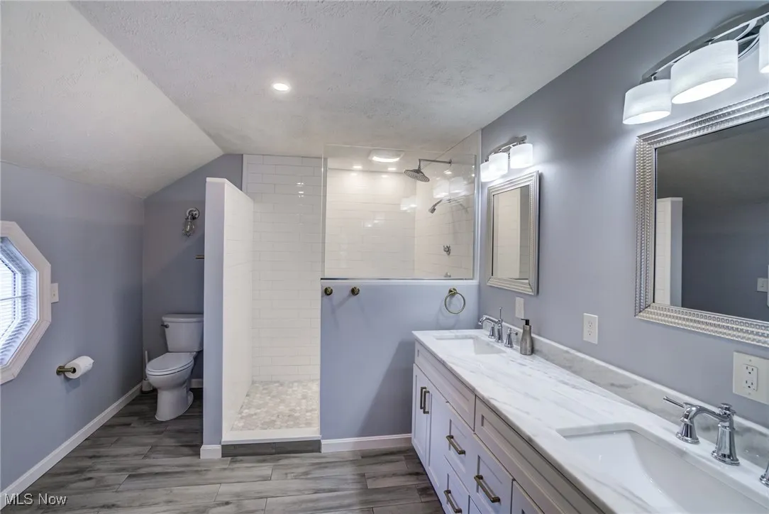 Full bath with double vanity, walk in shower, wood finished floors, a textured ceiling, and lofted ceiling