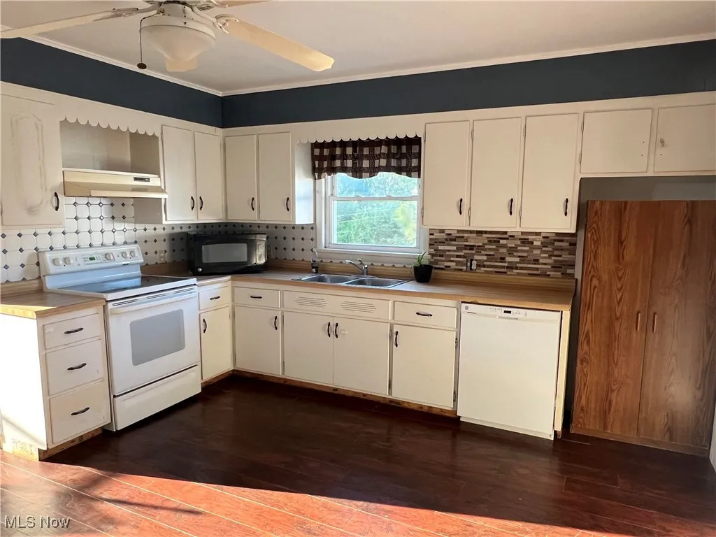 Kitchen featuring white appliances, dark wood-style flooring, backsplash, under cabinet range hood, and crown molding