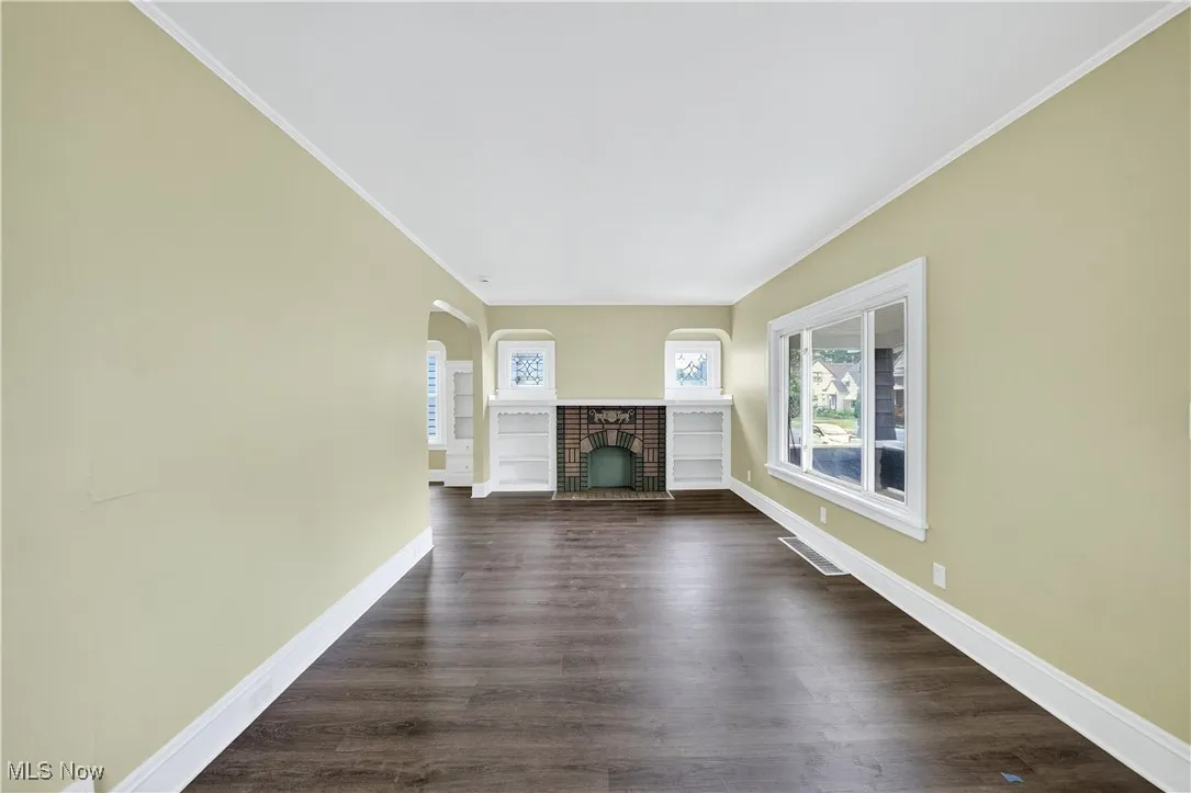 Unfurnished living room featuring a brick fireplace, ornamental molding, arched walkways, and dark wood finished floors