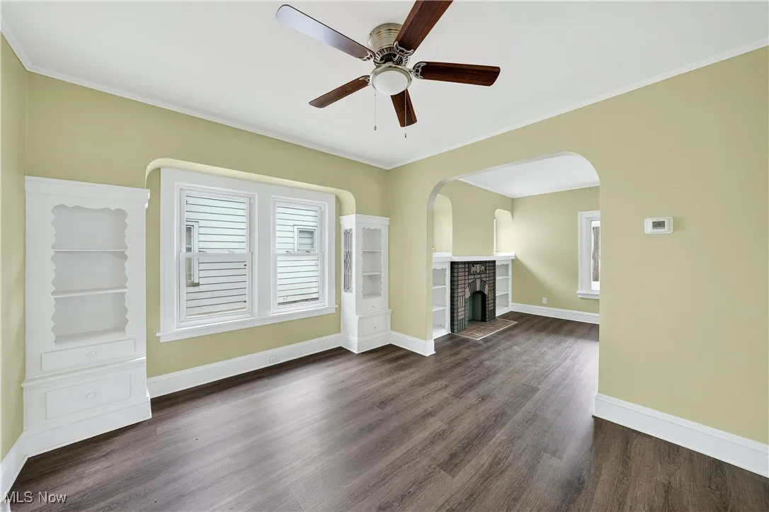 Unfurnished living room with dark wood-style floors, a ceiling fan, a brick fireplace, arched walkways, and ornamental molding