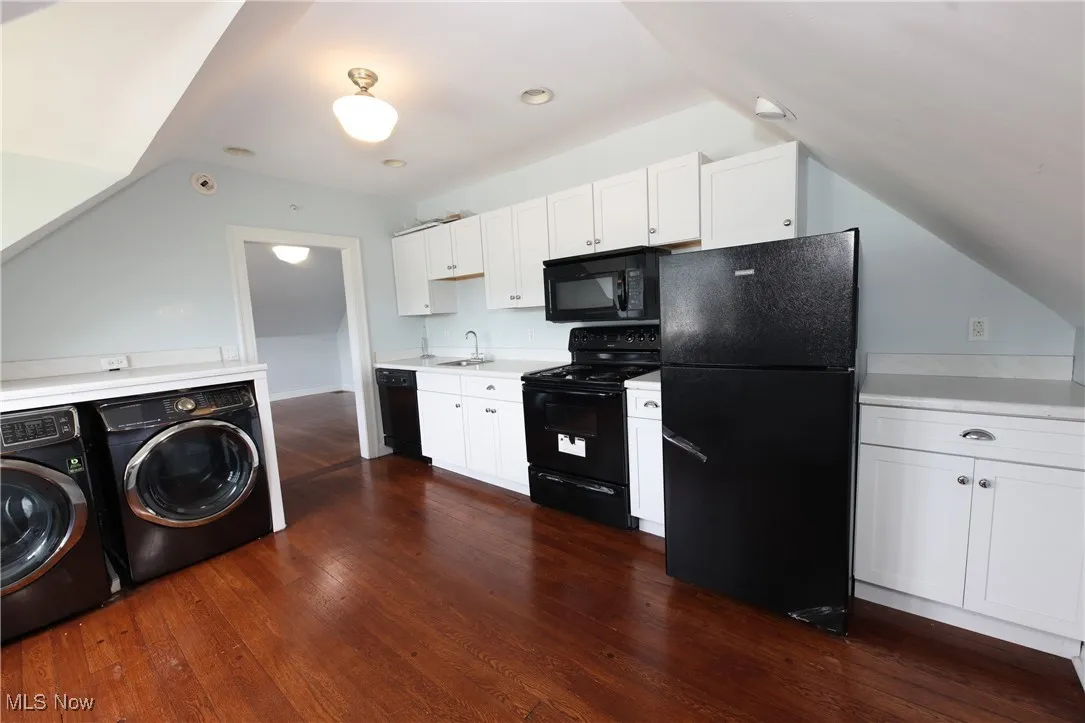 Kitchen with black appliances, washer and dryer, vaulted ceiling, white cabinetry, and light countertops