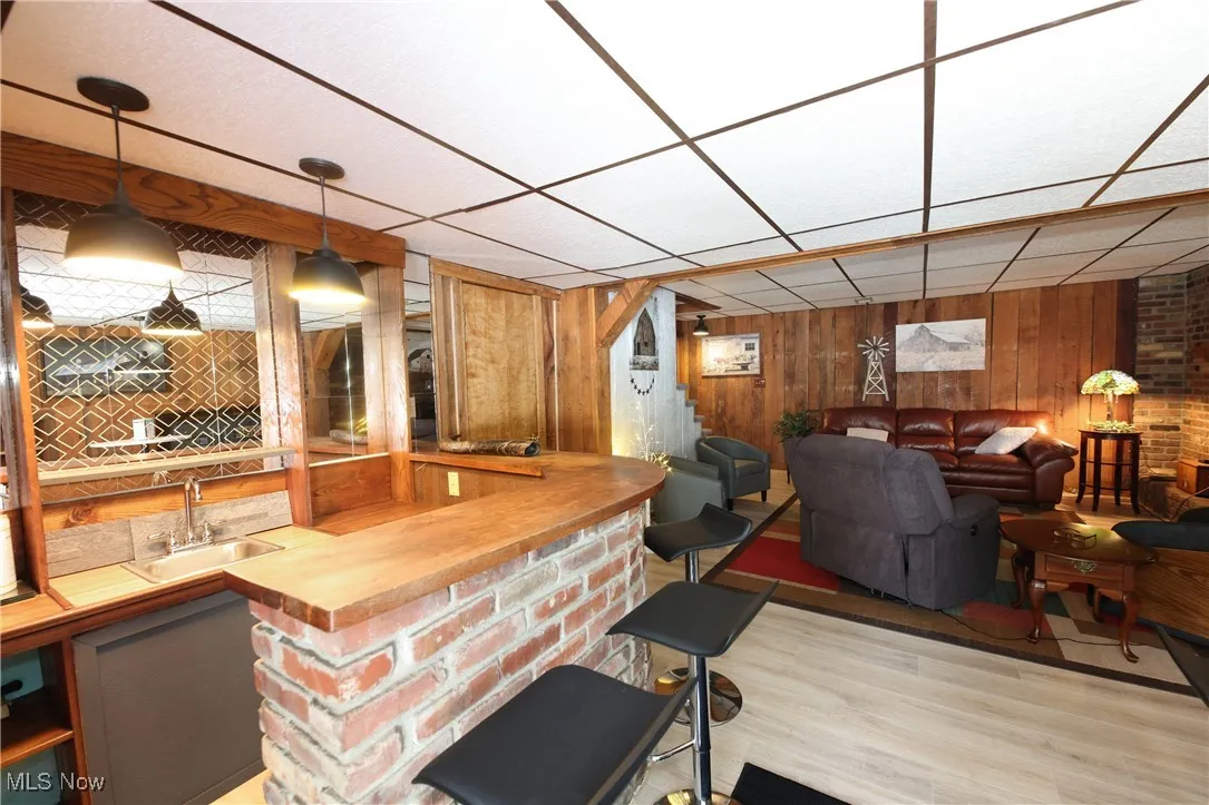 Indoor wet bar with wood walls, light wood-style flooring, hanging light fixtures, and a drop ceiling