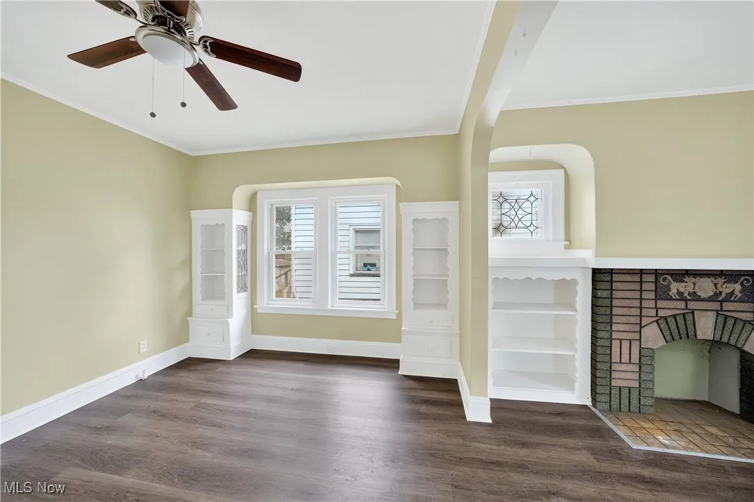 Unfurnished living room with plenty of natural light, wood finished floors, crown molding, a fireplace, and a ceiling fan