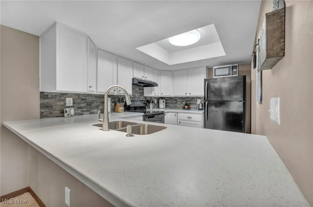 Kitchen with black appliances, under cabinet range hood, a raised ceiling, backsplash, and light countertops