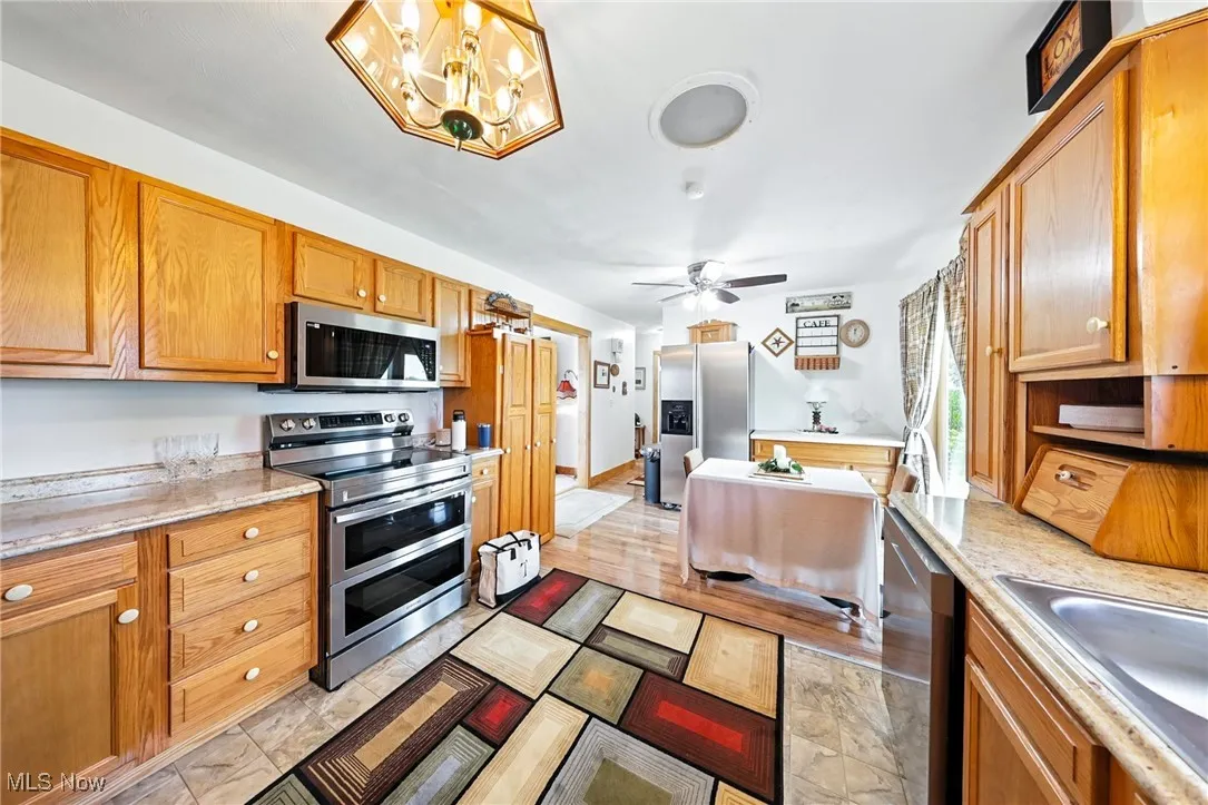 Kitchen featuring stainless steel appliances, ceiling fan, a chandelier, brown cabinets, and light stone counters