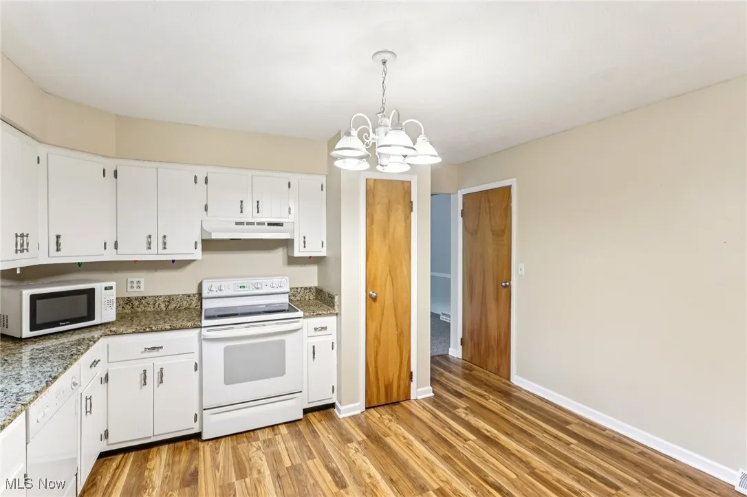 Kitchen with white appliances, under cabinet range hood, a chandelier, light wood-style floors, and white cabinetry