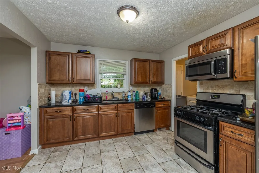 Kitchen featuring appliances with stainless steel finishes, backsplash, dark countertops, brown cabinets, and a textured ceiling