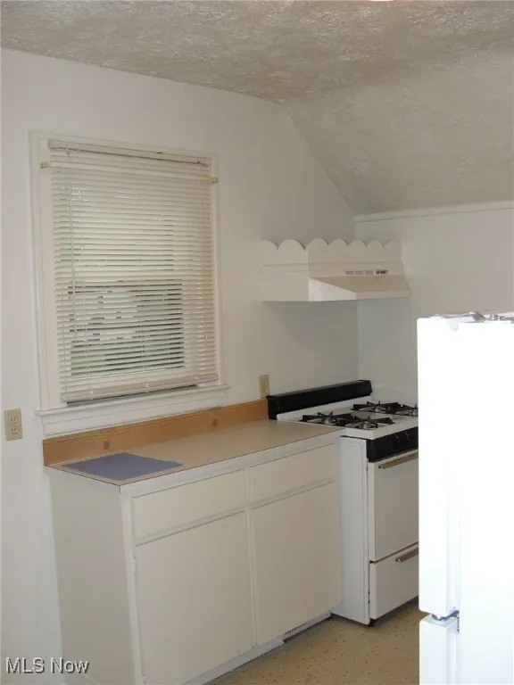 Kitchen featuring white appliances, a textured ceiling, lofted ceiling, under cabinet range hood, and white cabinets