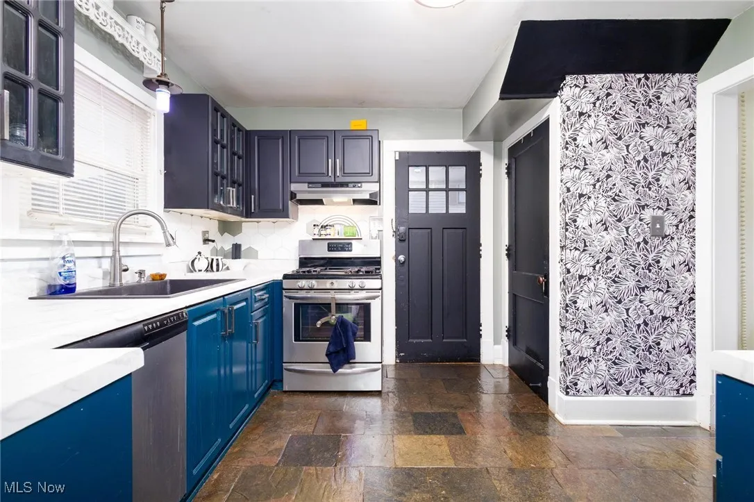 Kitchen featuring appliances with stainless steel finishes, under cabinet range hood, blue cabinetry, dark stone finish floors, and glass insert cabinets