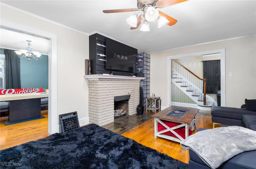 Living room featuring wood finished floors, a brick fireplace, a ceiling fan, stairway, and crown molding