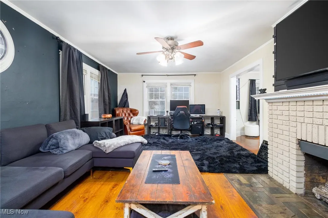 Living room with a ceiling fan, wood finished floors, an office area, ornamental molding, and a brick fireplace