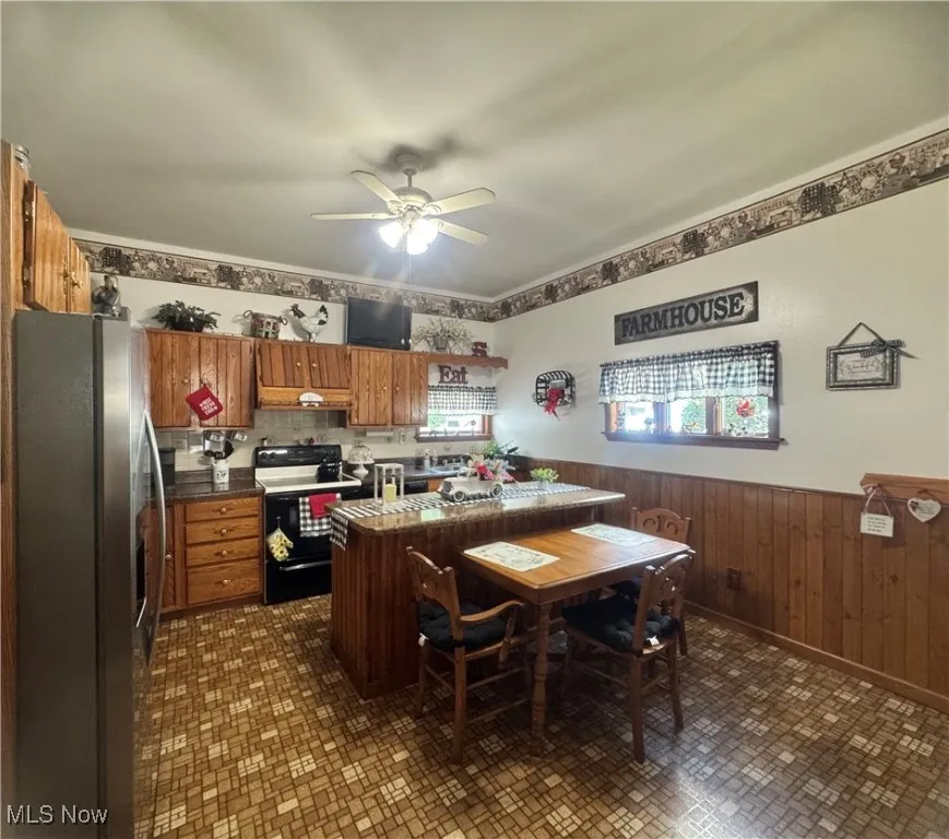 Kitchen with freestanding refrigerator, range with electric cooktop, wainscoting, brown cabinets, and ceiling fan