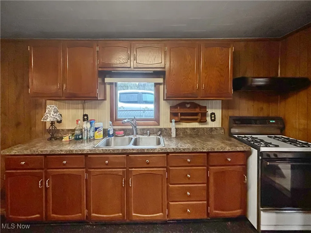 Kitchen featuring gas stove, exhaust hood, brown cabinetry, and wood walls