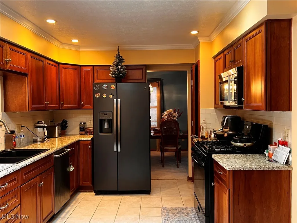 Kitchen featuring appliances with stainless steel finishes, decorative backsplash, light tile patterned flooring, light stone counters, and crown molding