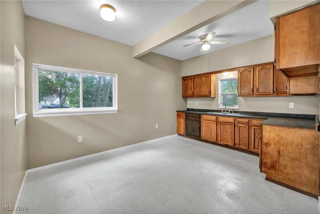 Kitchen with brown cabinets, a ceiling fan, dark countertops, beam ceiling, and light flooring