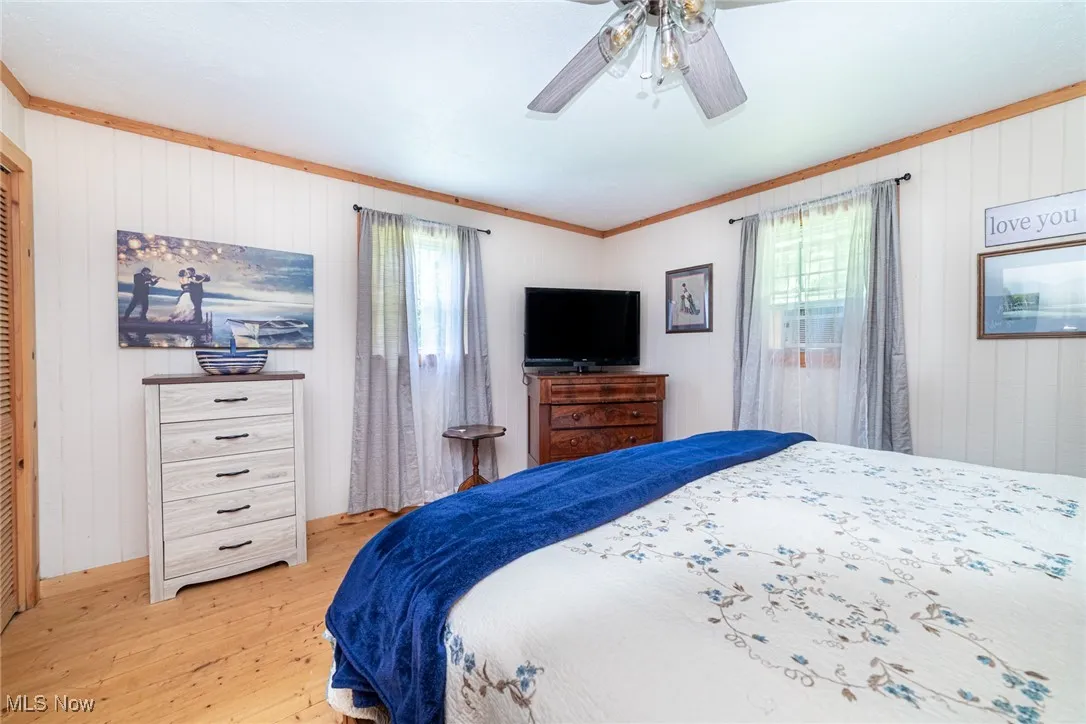 Bedroom with light wood-type flooring, multiple windows, a ceiling fan, wooden walls, and ornamental molding