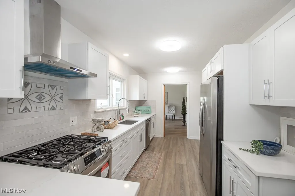 Kitchen with stainless steel appliances, wall chimney range hood, light wood-type flooring, and white cabinetry