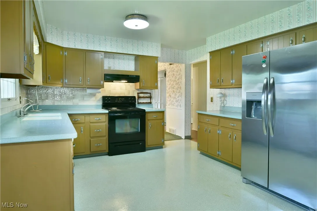 Kitchen with wallpapered walls, stainless steel refrigerator with ice dispenser, black electric range, light countertops, and backsplash