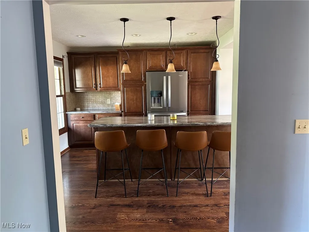 Kitchen with stainless steel fridge, dark wood-type flooring, a breakfast bar, backsplash, and decorative light fixtures