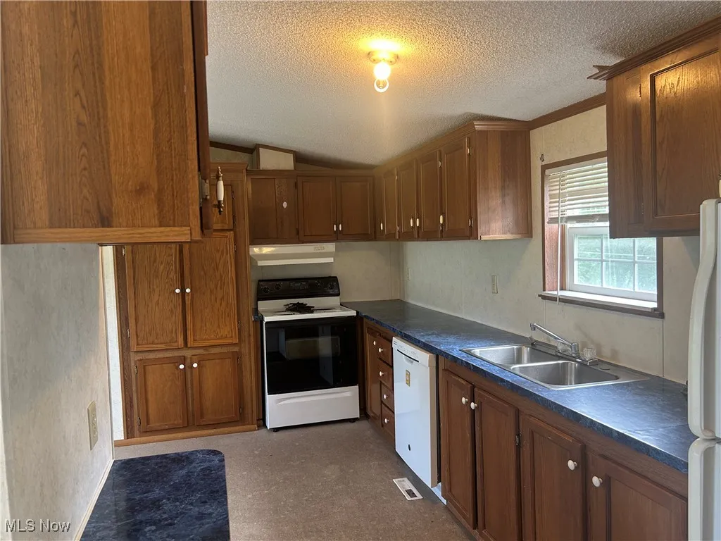 Kitchen with white appliances, a textured ceiling, brown cabinetry, under cabinet range hood, and dark countertops
