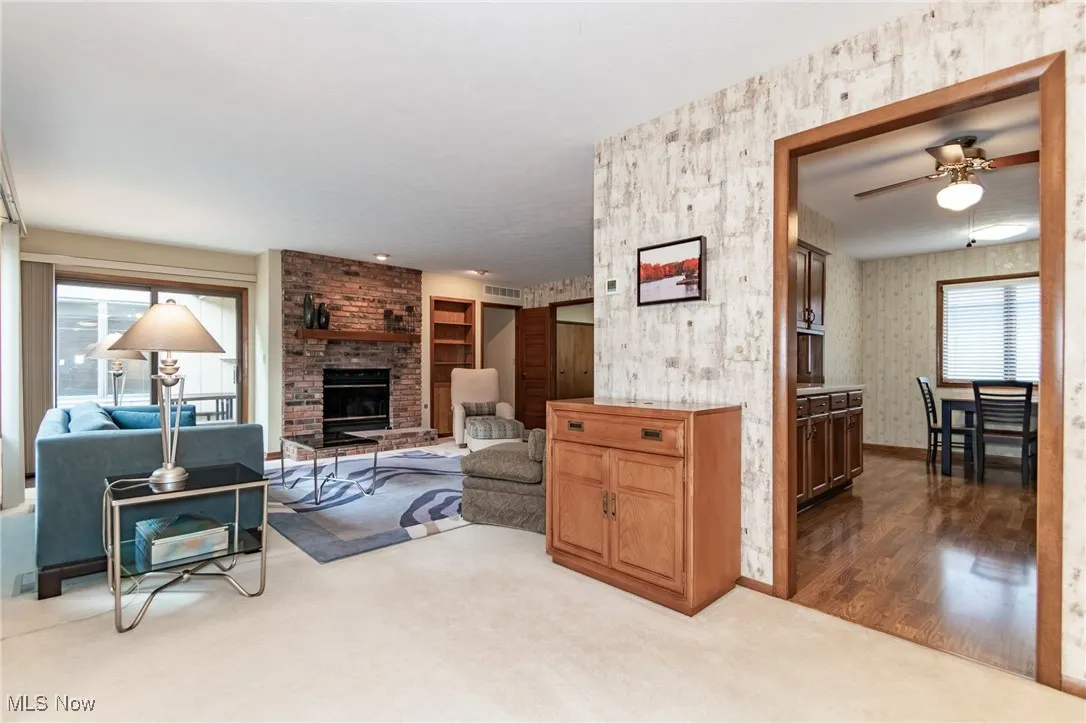 Carpeted living area with wallpapered walls, a brick fireplace, and a ceiling fan