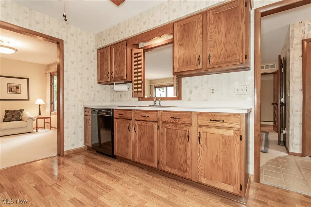 Kitchen with wallpapered walls, light wood finished floors, light countertops, black dishwasher, and brown cabinets