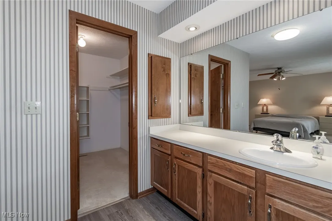 Ensuite bathroom featuring wallpapered walls, wood finished floors, vanity, and a ceiling fan