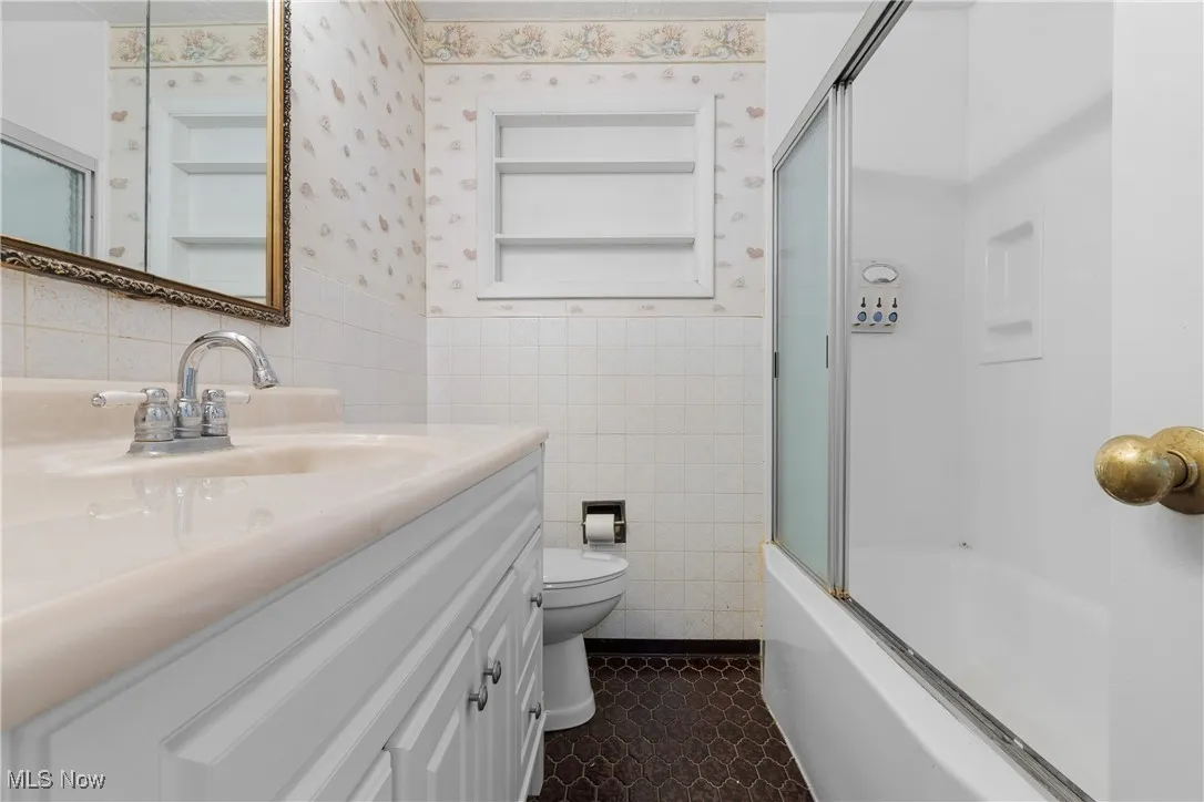 Full bathroom with tile walls, vanity, combined bath / shower with glass door, a wainscoted wall, and wallpapered walls