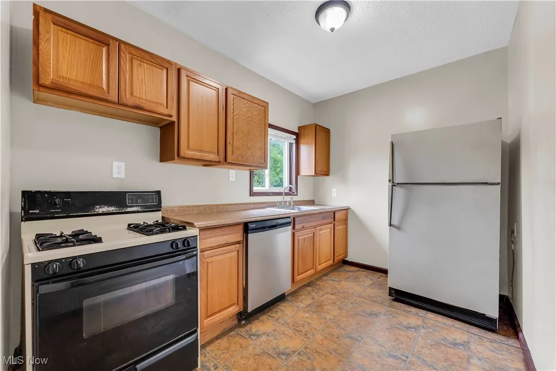 Kitchen featuring black gas stove, freestanding refrigerator, stainless steel dishwasher, light countertops, and light stone finish floors