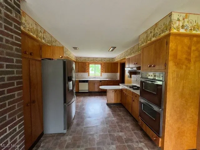 Kitchen with a peninsula, stainless steel appliances, brown cabinetry, light countertops, and backsplash