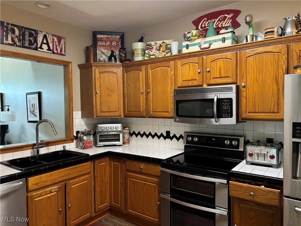 Kitchen with tile counters, appliances with stainless steel finishes, decorative backsplash, and brown cabinets