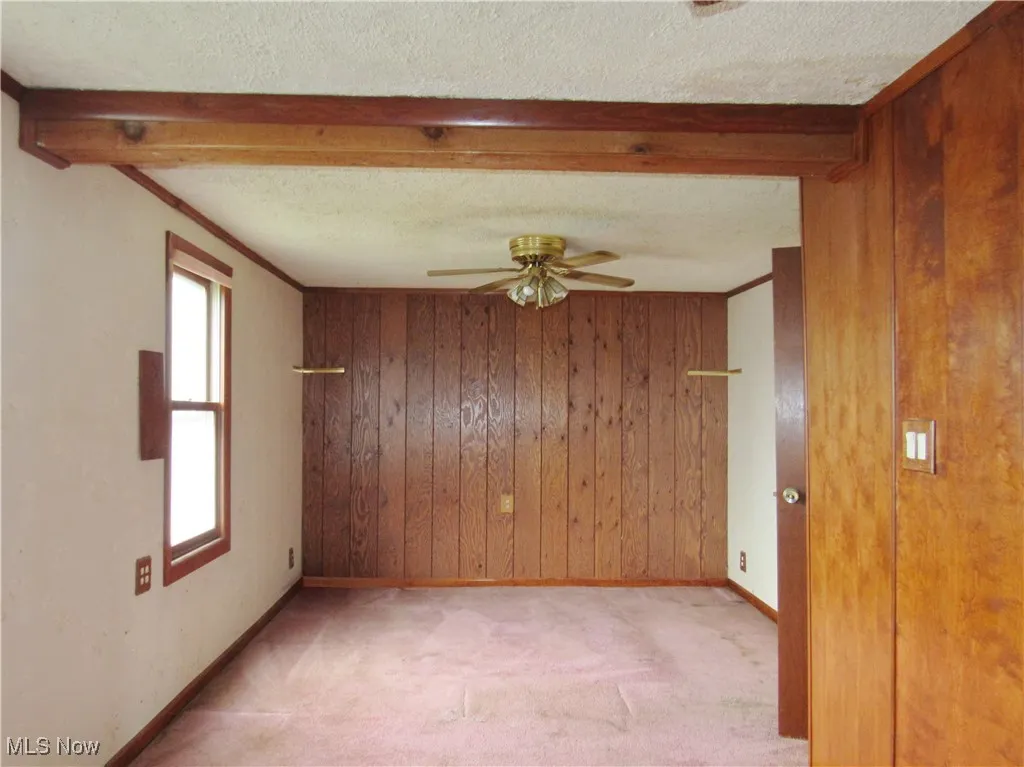 Unfurnished room featuring ceiling fan, light colored carpet, beamed ceiling, wooden walls, and a textured ceiling
