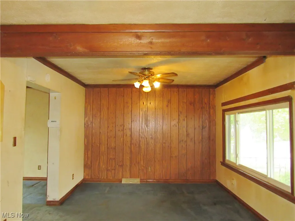 Empty room featuring carpet, beamed ceiling, wood walls, a ceiling fan, and ornamental molding