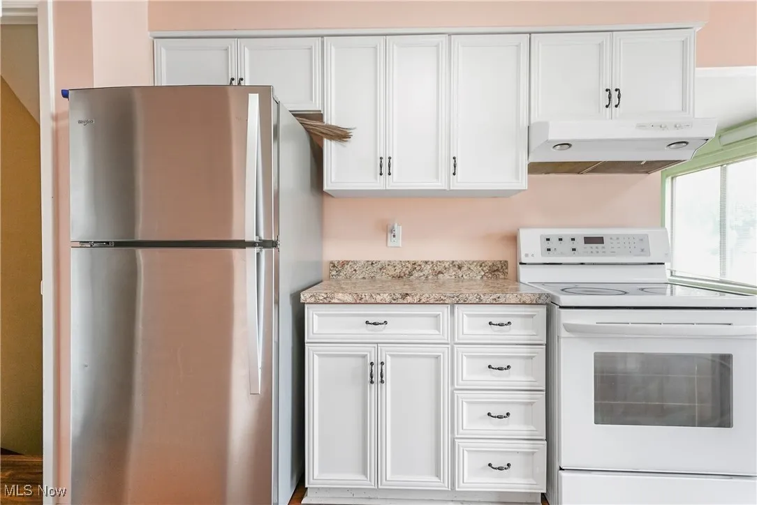 Kitchen featuring white electric range, freestanding refrigerator, under cabinet range hood, white cabinets, and light stone countertops