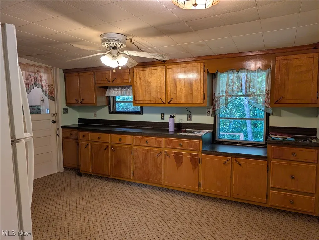 Kitchen with brown cabinets, freestanding refrigerator, dark countertops, and a ceiling fan