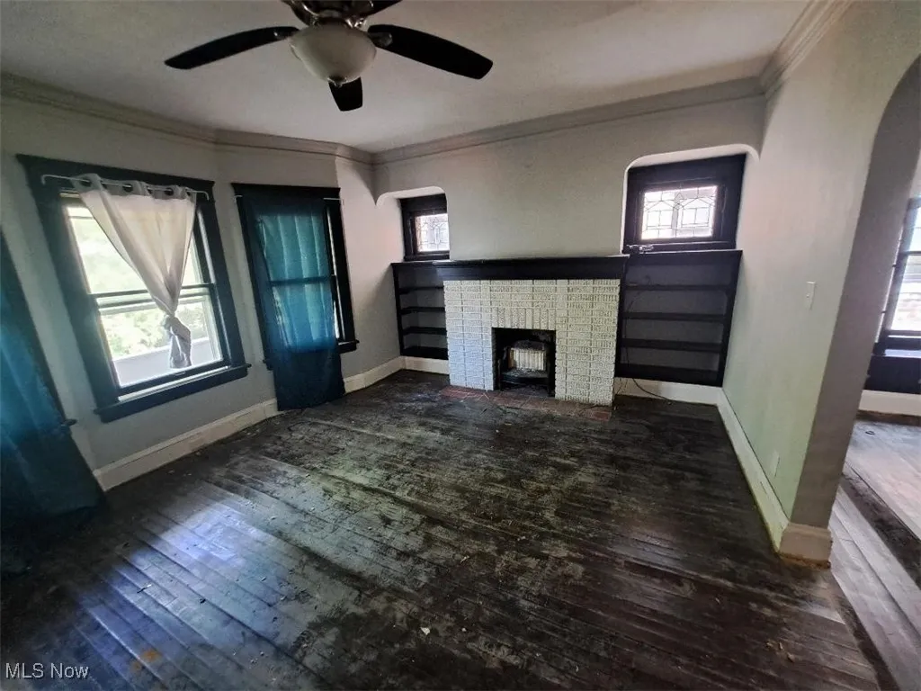 Unfurnished living room featuring hardwood / wood-style flooring, a brick fireplace, crown molding, arched walkways, and ceiling fan