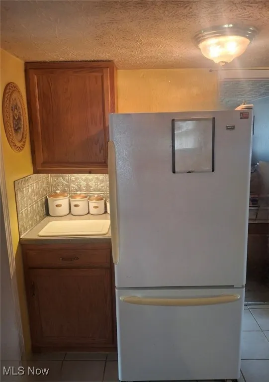 Kitchen featuring freestanding refrigerator, light tile patterned floors, brown cabinets, and decorative backsplash