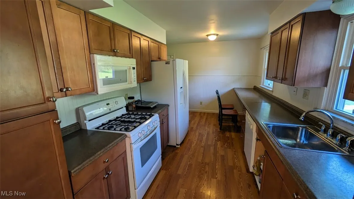 Kitchen with white appliances, dark wood-style flooring, dark countertops, and brown cabinets