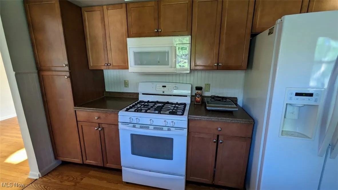 Kitchen featuring white appliances, dark countertops, dark wood-style floors, and brown cabinets