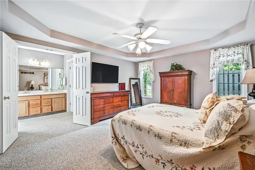 Carpeted bedroom with a tray ceiling, multiple windows, connected bathroom, and ceiling fan