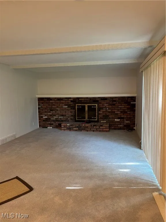 Unfurnished living room featuring carpet floors, beamed ceiling, and brick wall