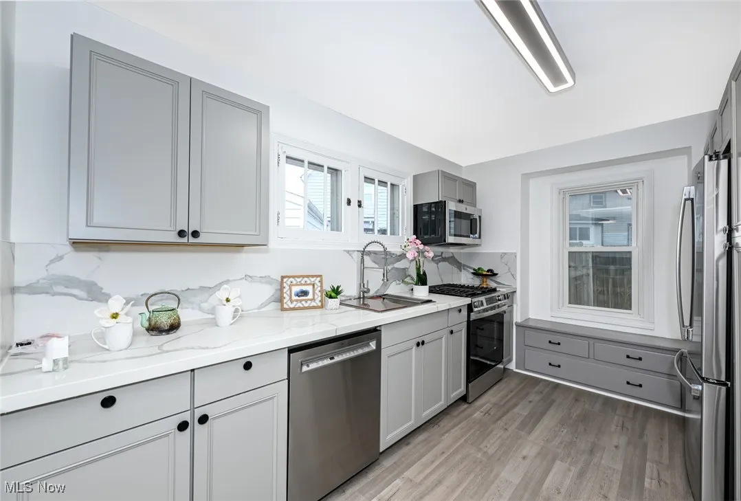 Kitchen with appliances with stainless steel finishes, gray cabinetry, light wood-type flooring, light stone countertops, and backsplash