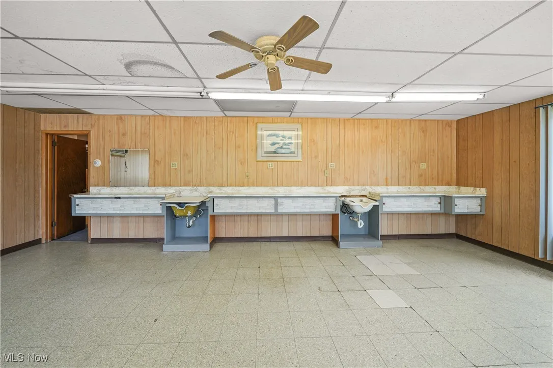Miscellaneous room with light floors, a paneled ceiling, wooden walls, a ceiling fan, and an office area