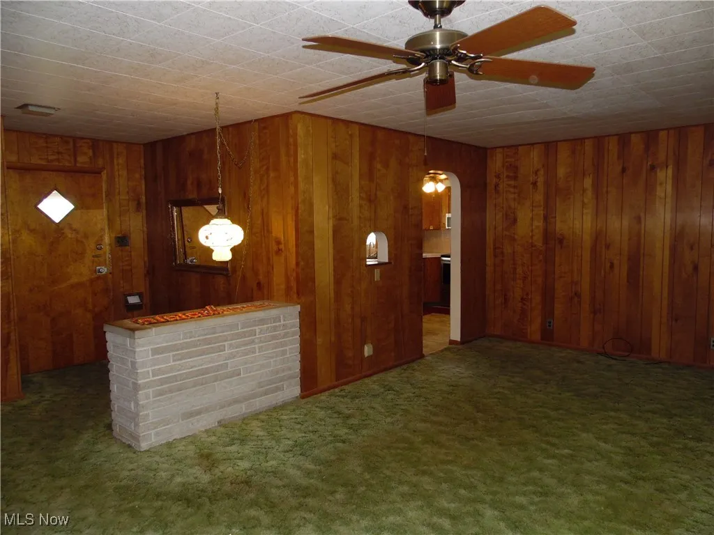 Unfurnished living room featuring carpet floors, wood walls, ceiling fan, and arched walkways