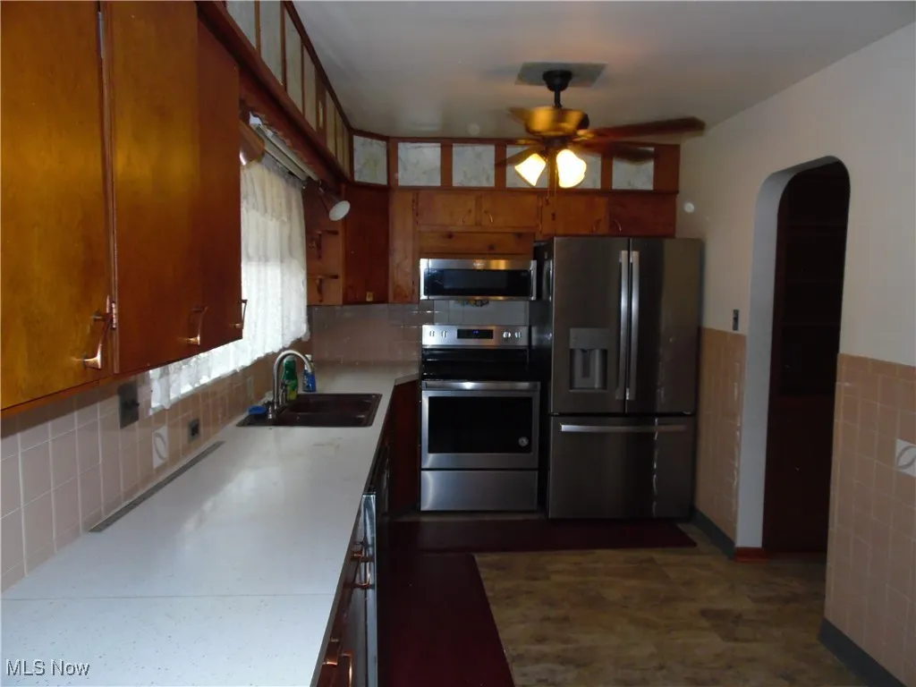 Kitchen with stainless steel appliances, ceiling fan, arched walkways, brown cabinetry, and tile walls