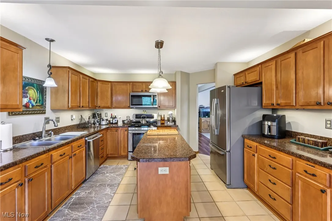 Kitchen with a sink, appliances, a kitchen island, brown cabinetry, pendant lighting, and ceramic flooring